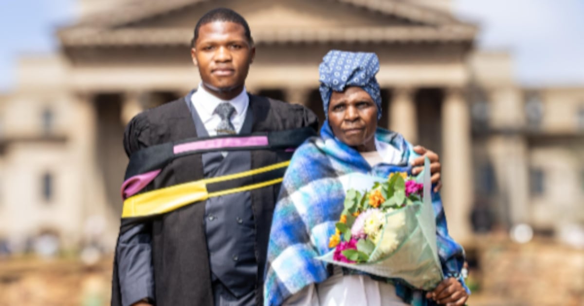 A graduate in a gown standing with an older woman holding flowers